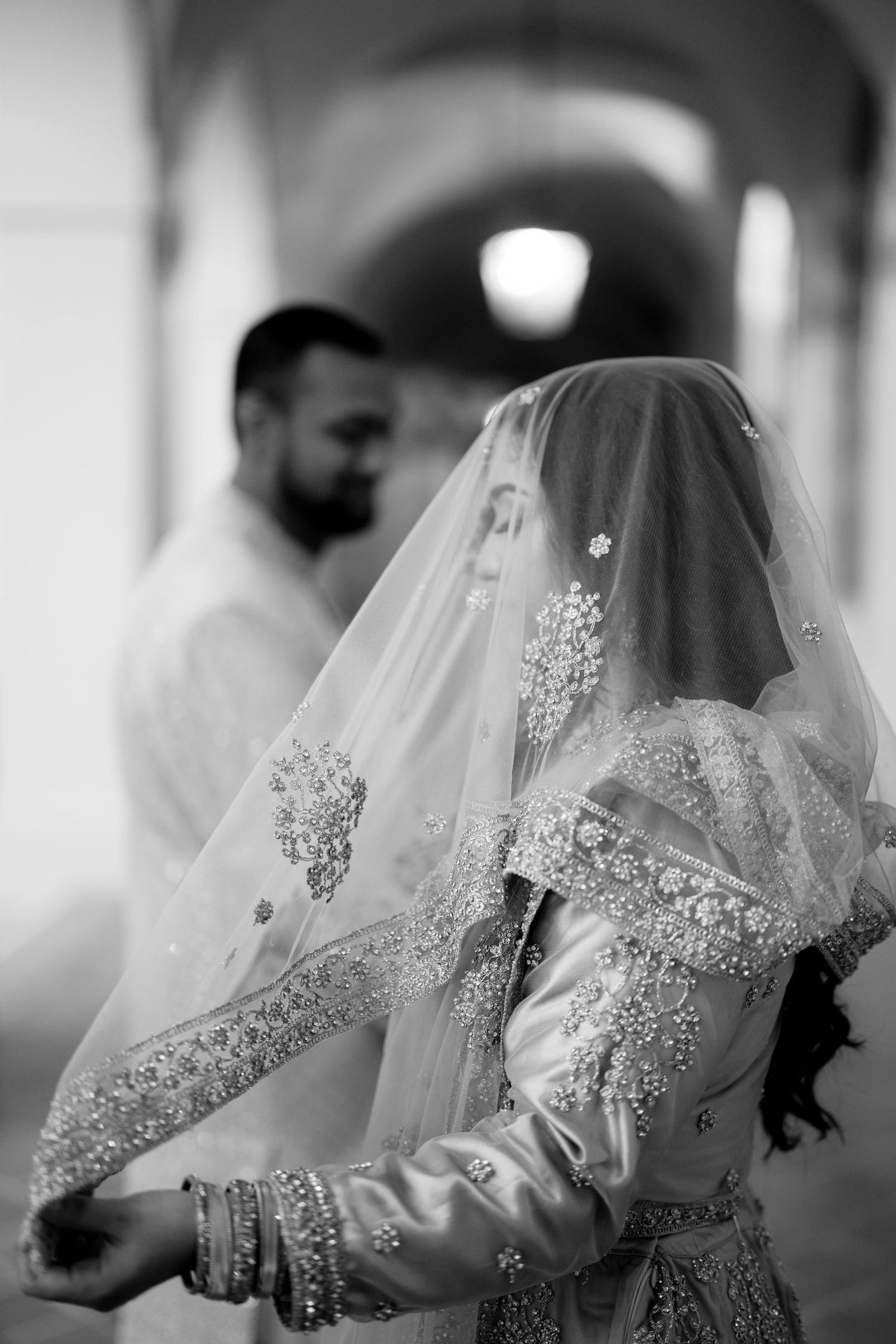 A black and white photo of a couple in traditional wedding attire under an arched hallway.