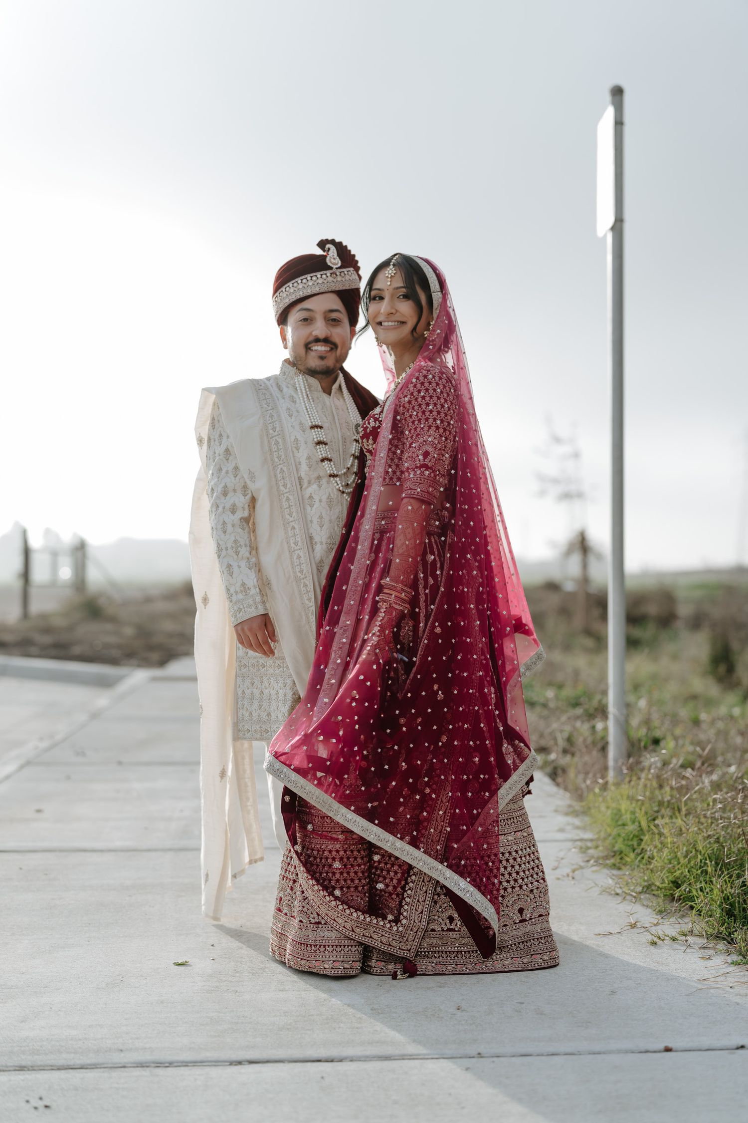 Couple in traditional Indian wedding attire posing outdoors on a concrete pathway.