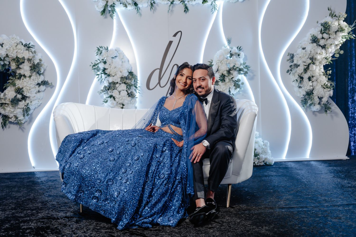 Couple seated on elegant white sofa against illuminated wavy wall with floral decorations.