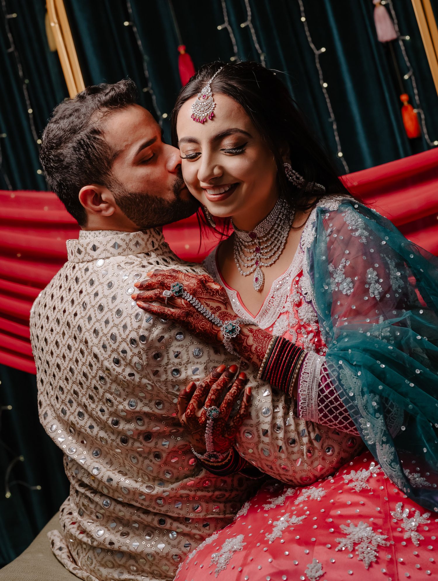 A romantic moment captured between a couple in traditional Indian wedding attire with sparkly backdrop and red decor.