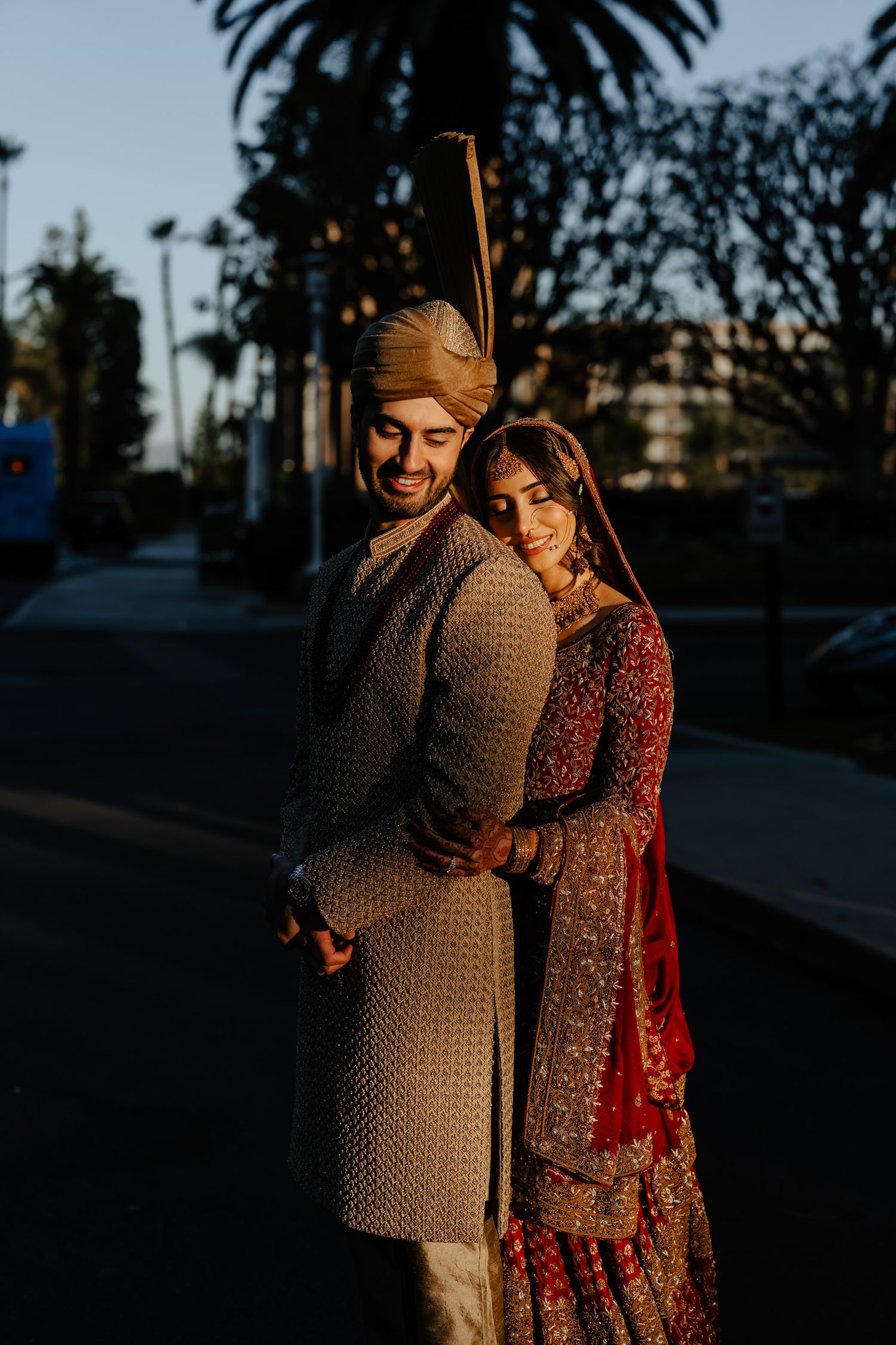 A couple in traditional South Asian wedding attire embrace during a sunset photoshoot outdoors.