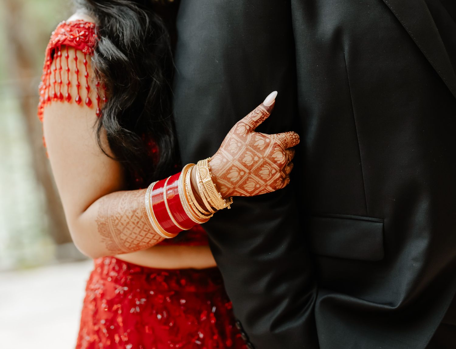 Close-up of hands with henna and red bangles against black formal wear.