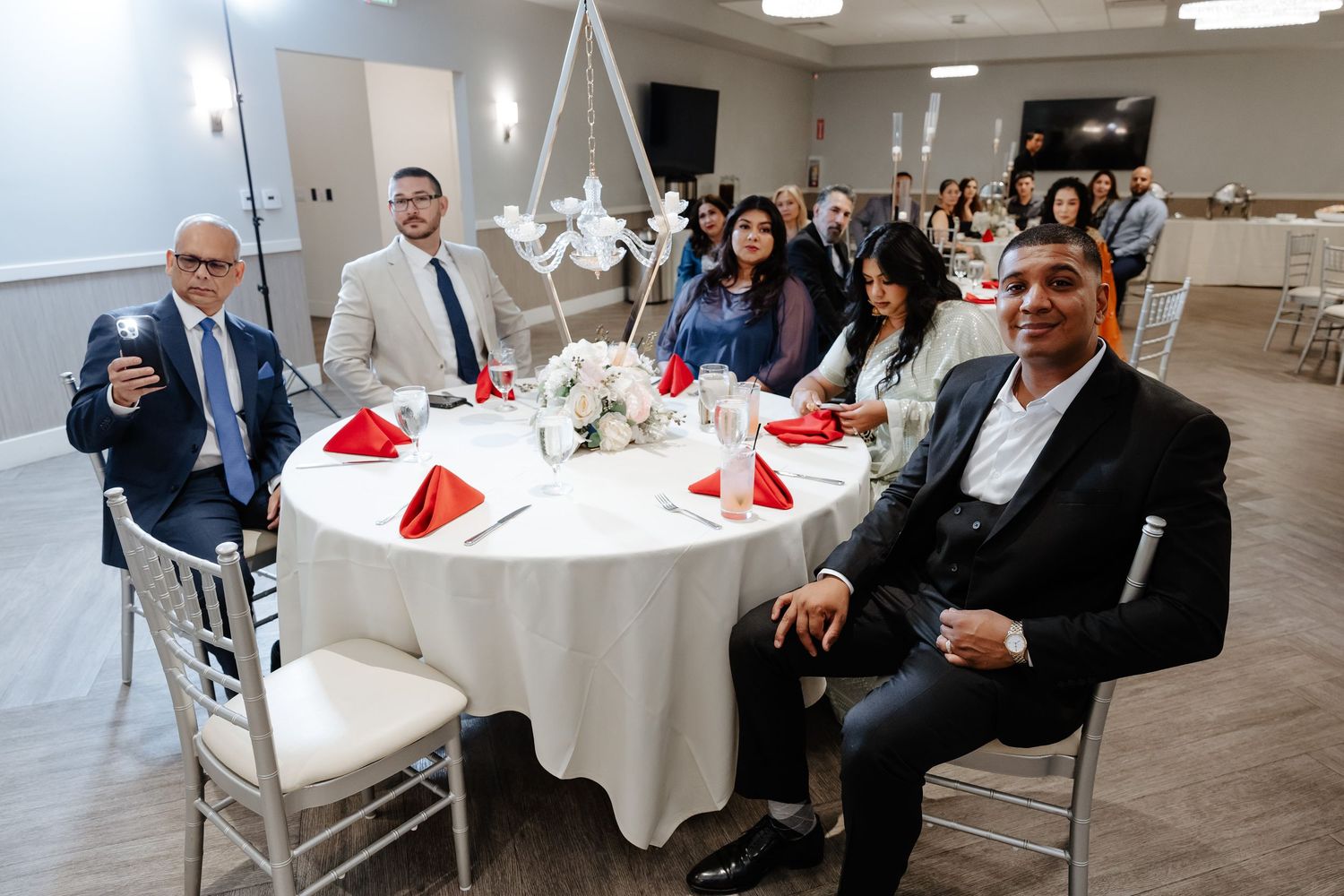 Group of elegantly dressed guests smiling at a decorated banquet table with red napkins at a formal event.