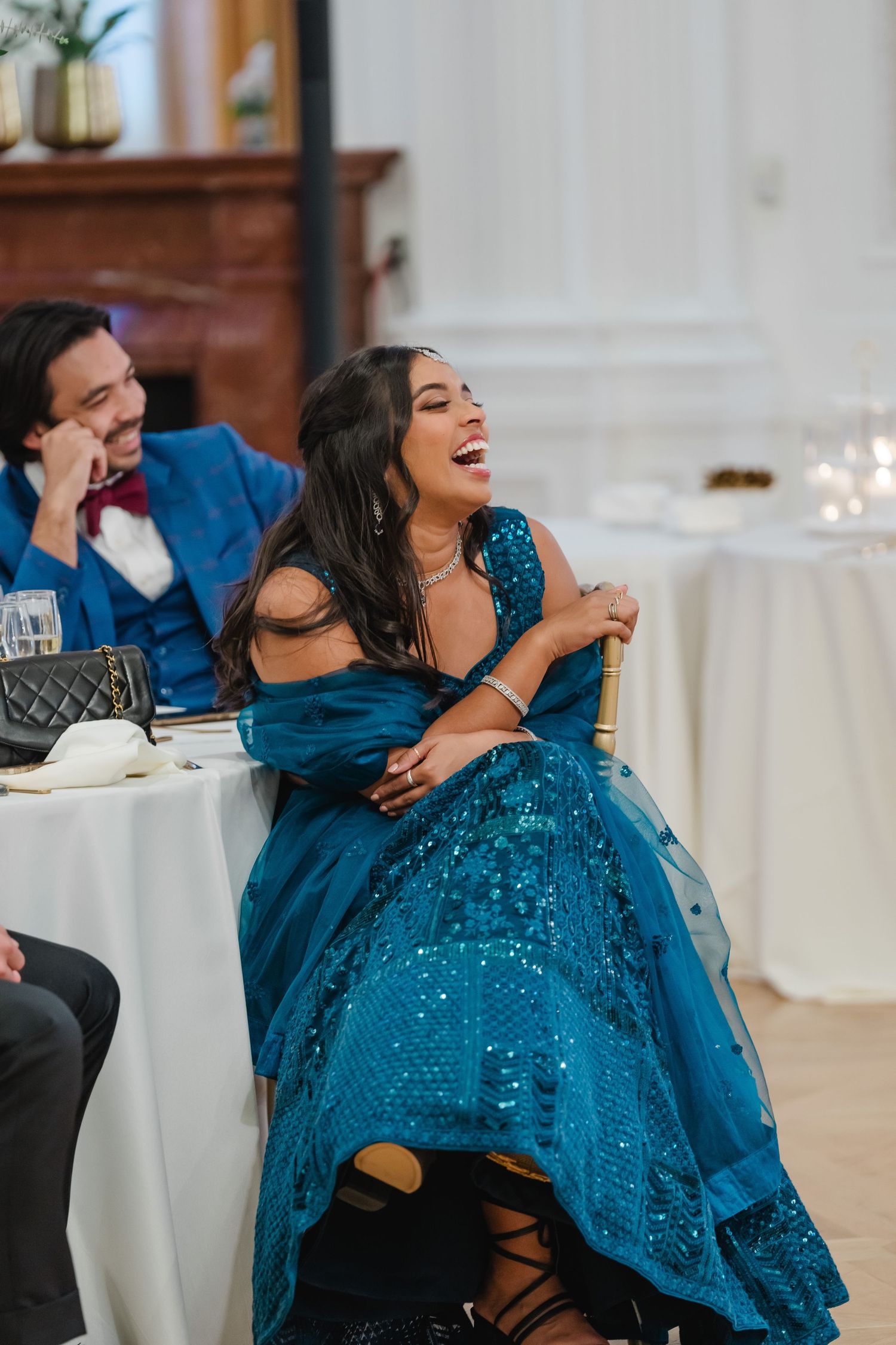 Two guests share a joyful moment of laughter at an elegant reception, with one wearing a sparkling teal evening gown.