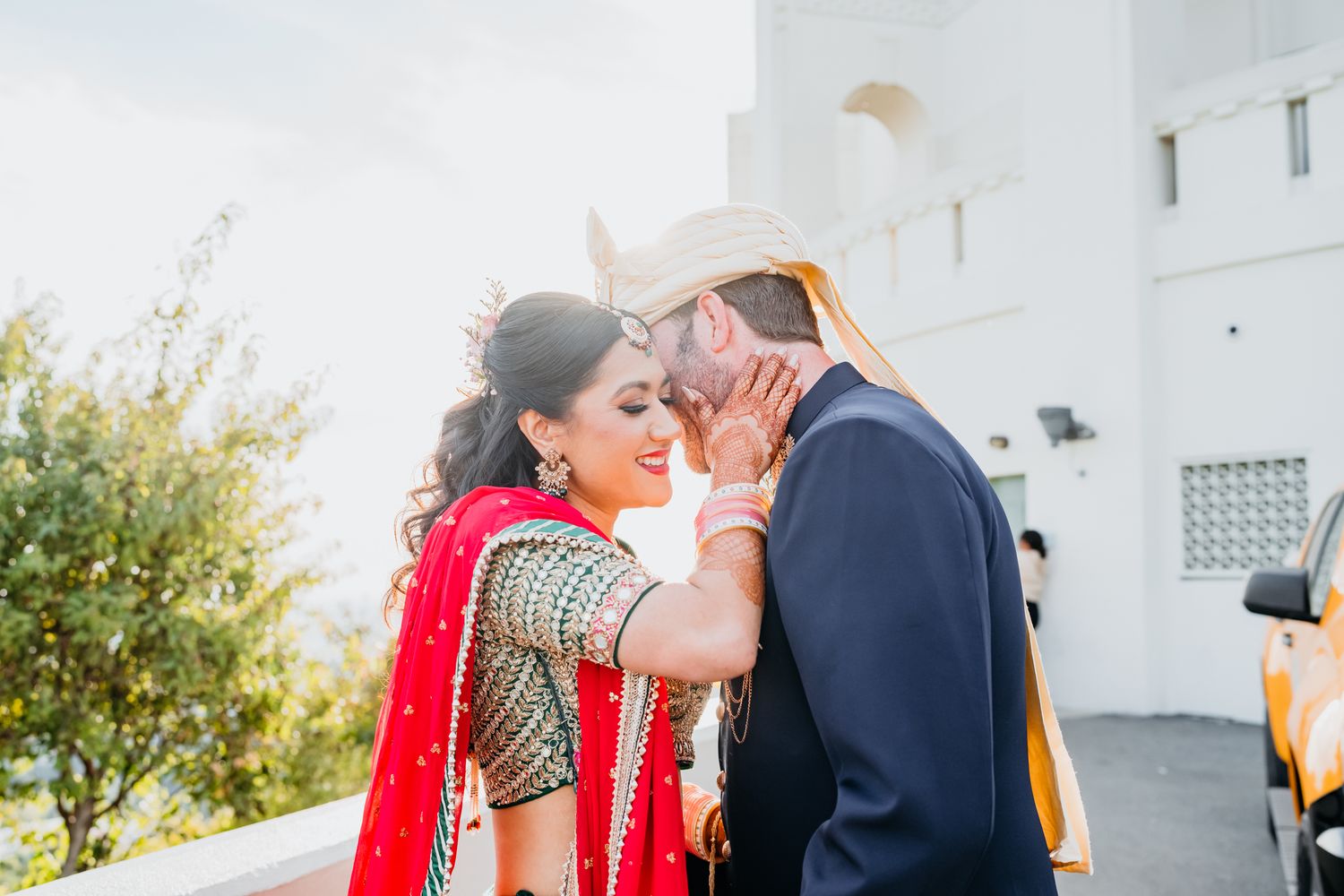 A couple in traditional South Asian wedding attire share an intimate moment outside a white building.