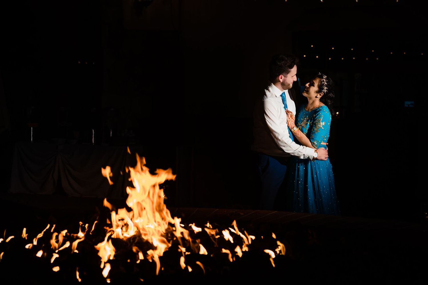A couple dances together at night with a dramatic fire display burning in the background.
