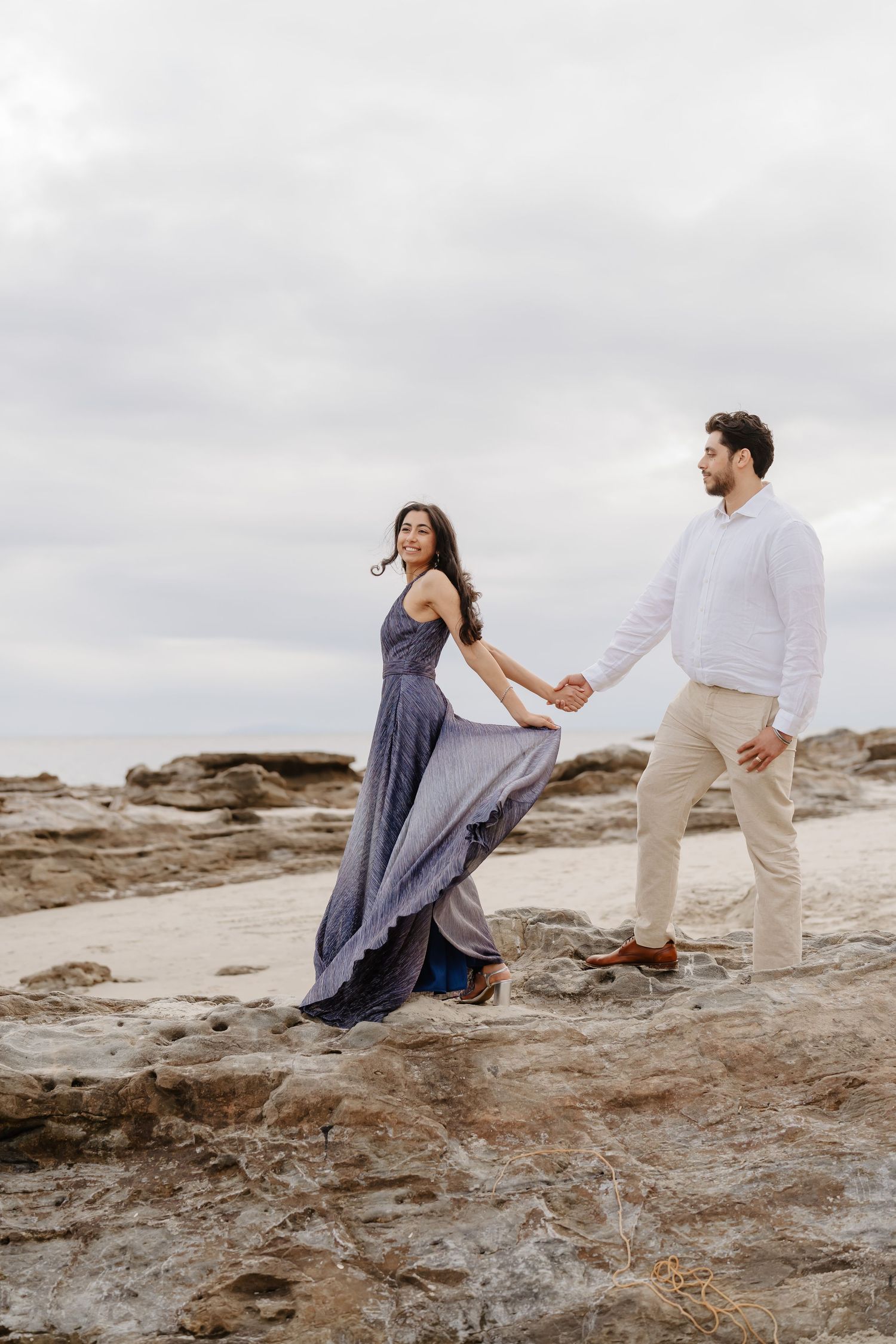Couple holding hands on rocky beach, woman in flowing blue dress, man in white shirt and khaki pants, overcast sky.
