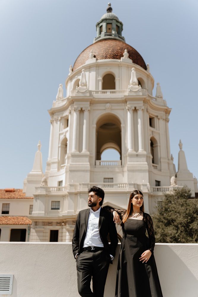 Elegant couple in black attire posing before Pasadena City Hall's iconic dome on a sunny day.