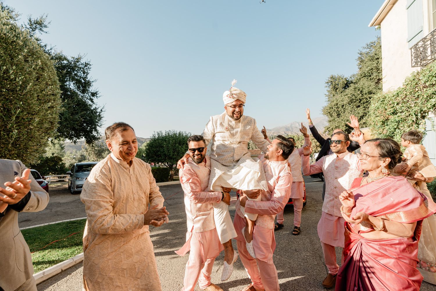 Wedding party members in pink and beige outfits celebrate joyfully outdoors during a summer celebration.