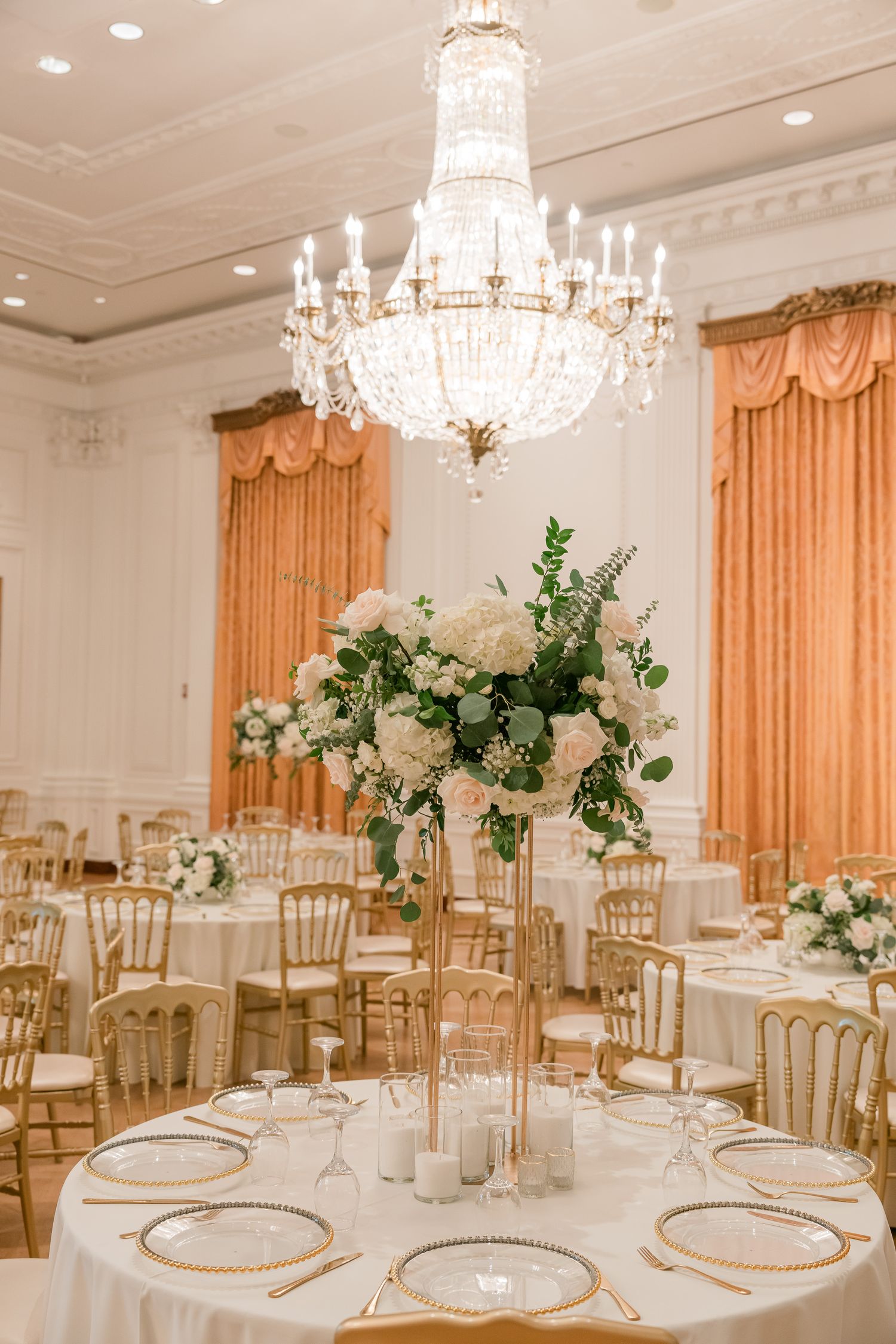 An elegant wedding reception room with crystal chandeliers and peach drapes features floral centerpieces on round tables.