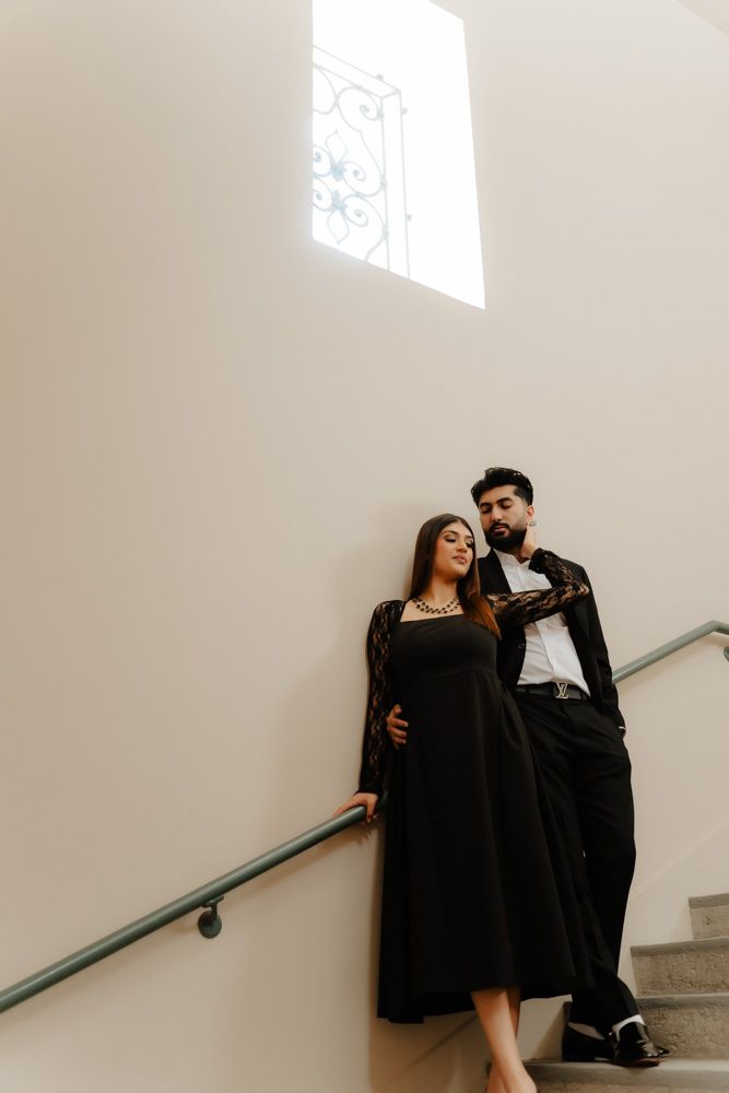Stylish couple in black outfits posing on staircase against a bright white wall with window.