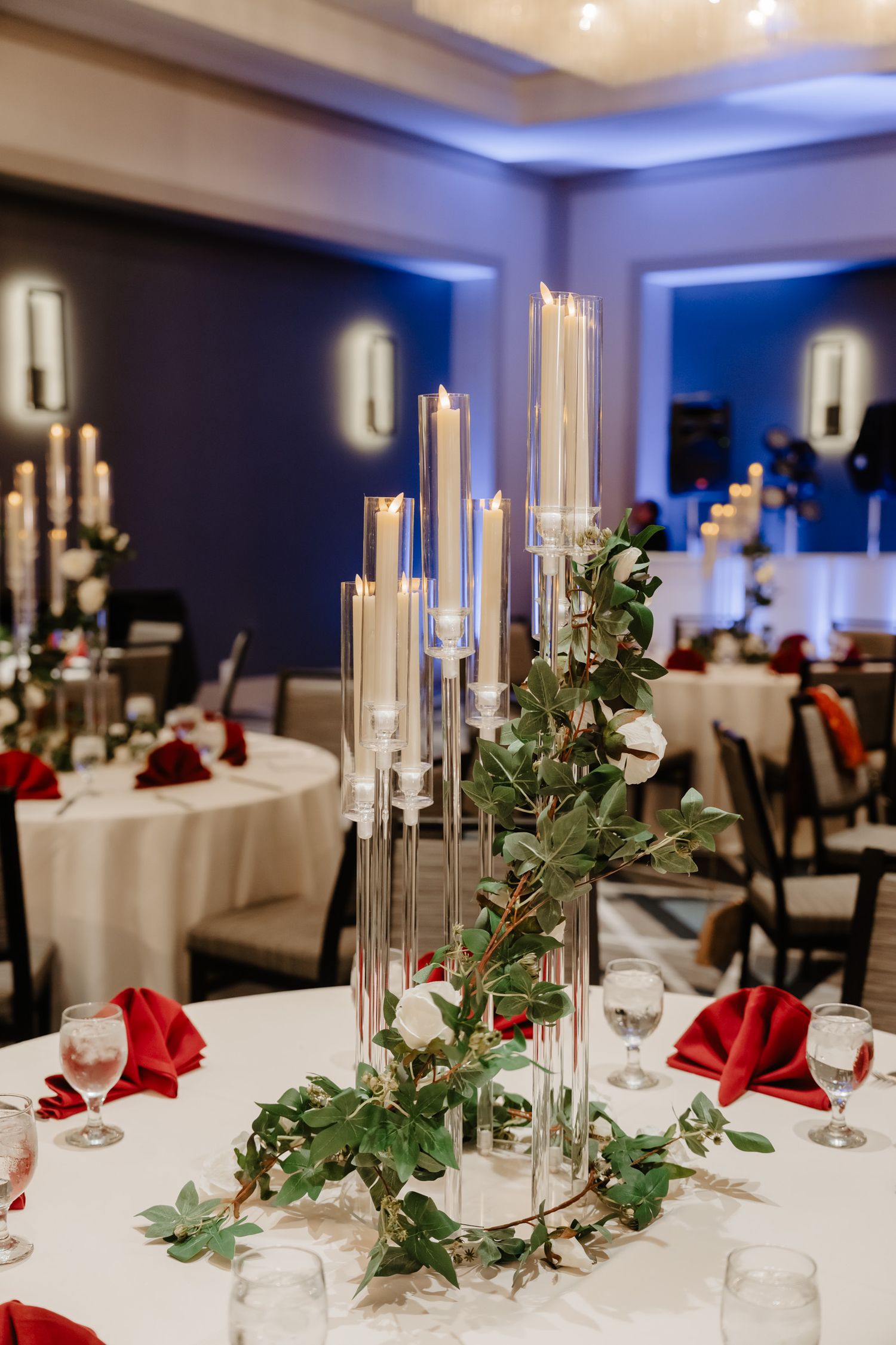 Elegant wedding reception tables with white linens, tall candles, and red napkins in a blue-lit banquet room.