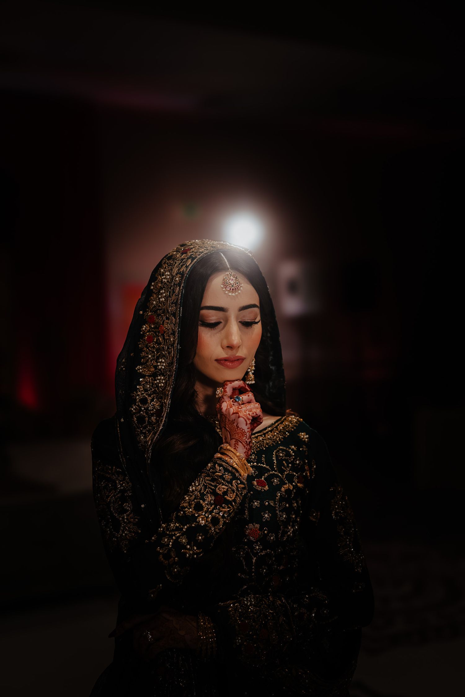 Moody portrait of someone in traditional South Asian bridal attire with henna-adorned hands and ornate embroidered dress.