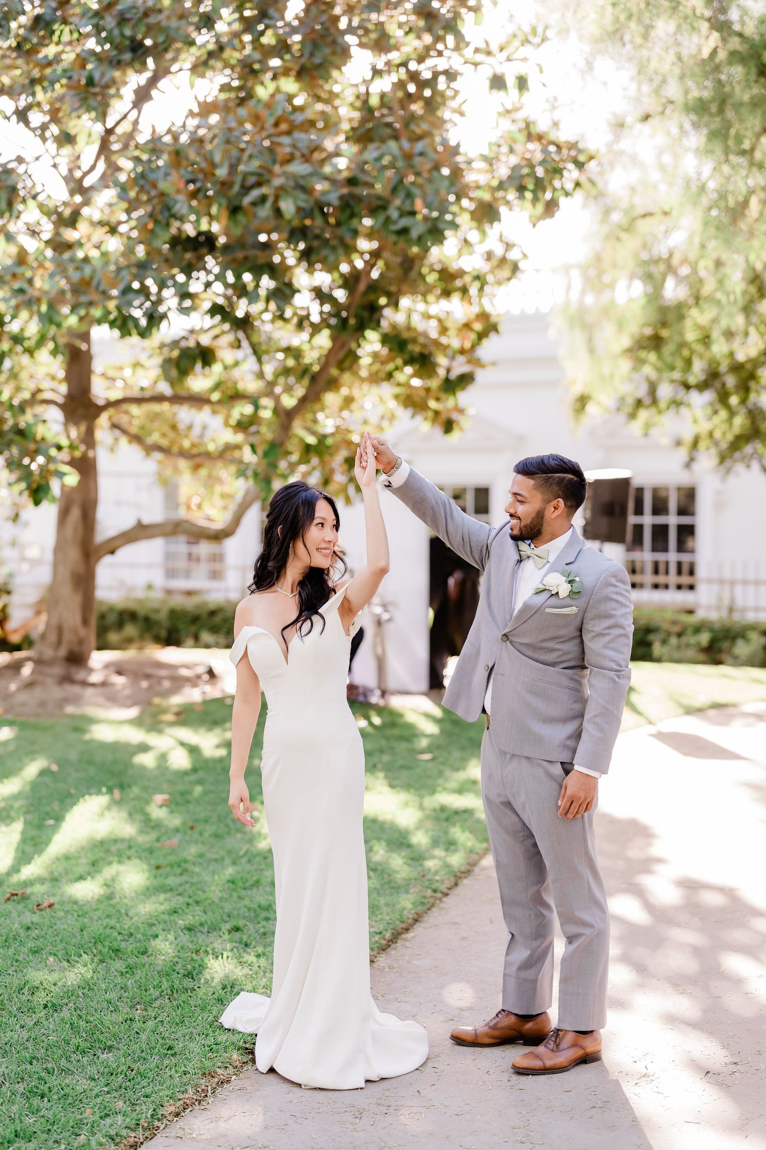 Newlywed couple in white dress and gray suit dance playfully together on a sunny garden path.
