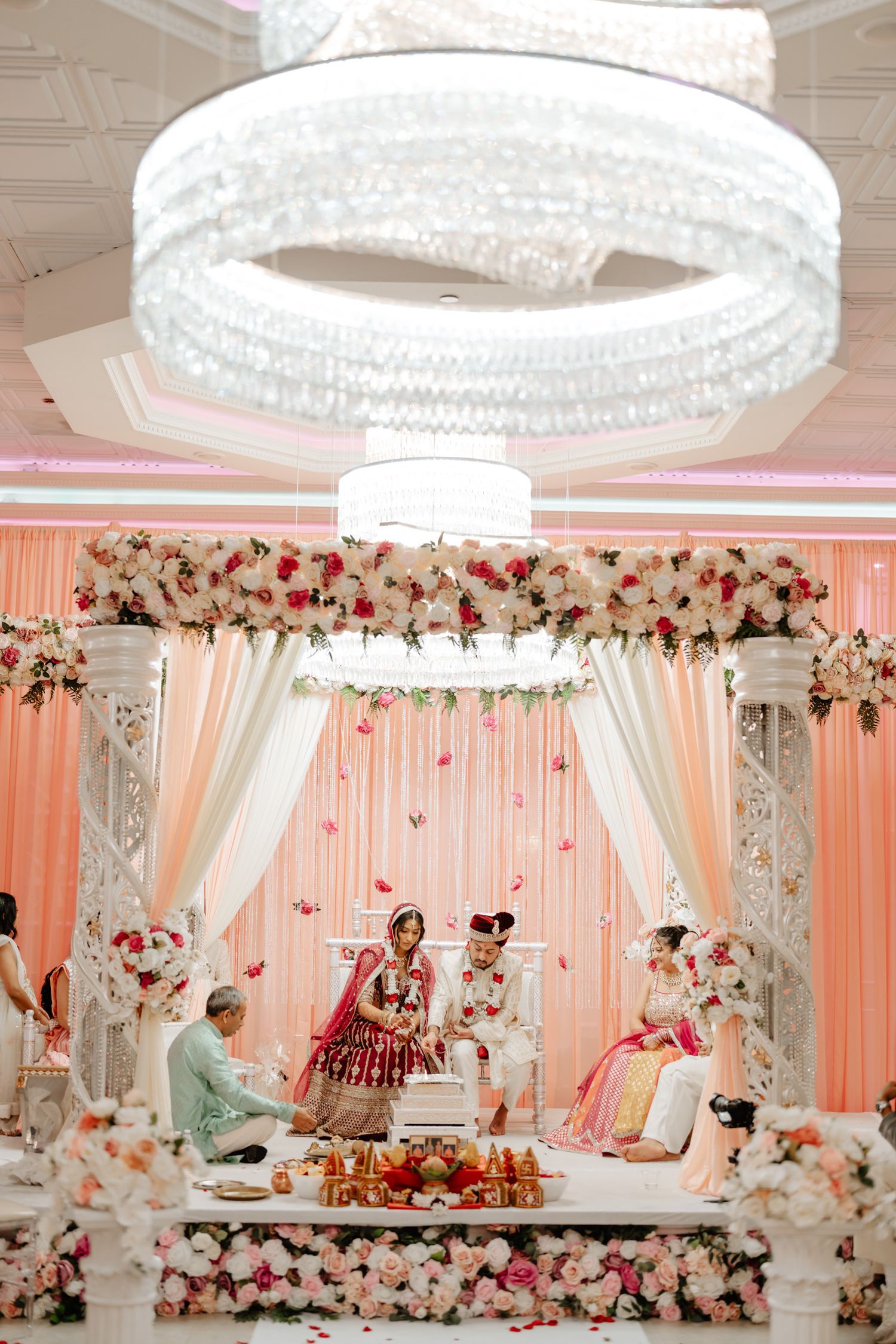 Elegant wedding mandap decorated with pink and white floral arrangements under crystal chandeliers.