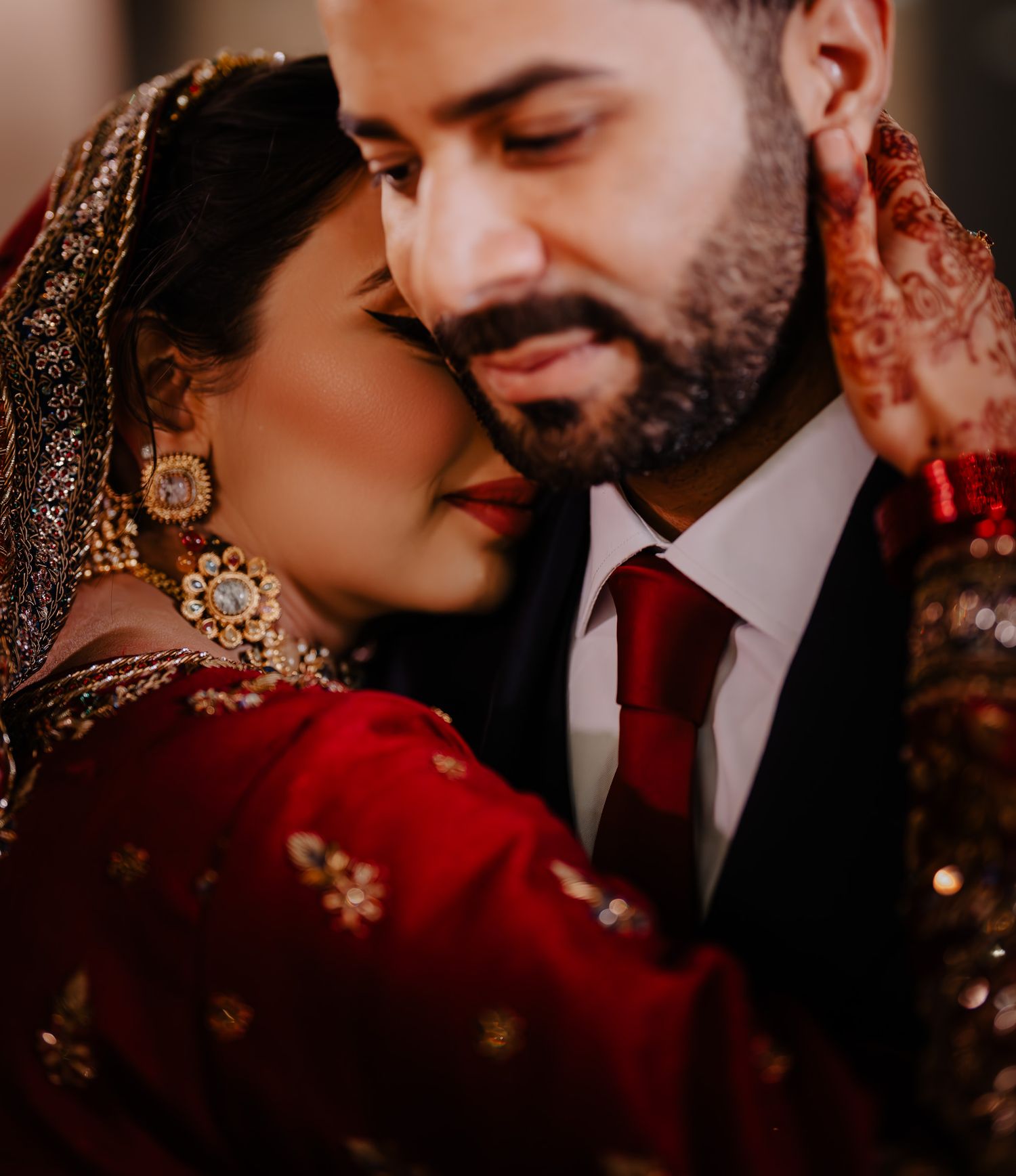 A romantic couple shares an intimate moment in traditional South Asian wedding attire with red and gold details.