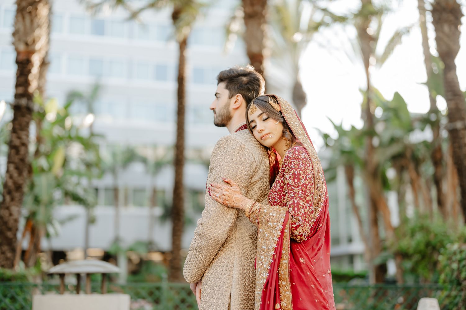 A couple in traditional Indian wedding attire embraces in a tropical garden setting with palm trees.