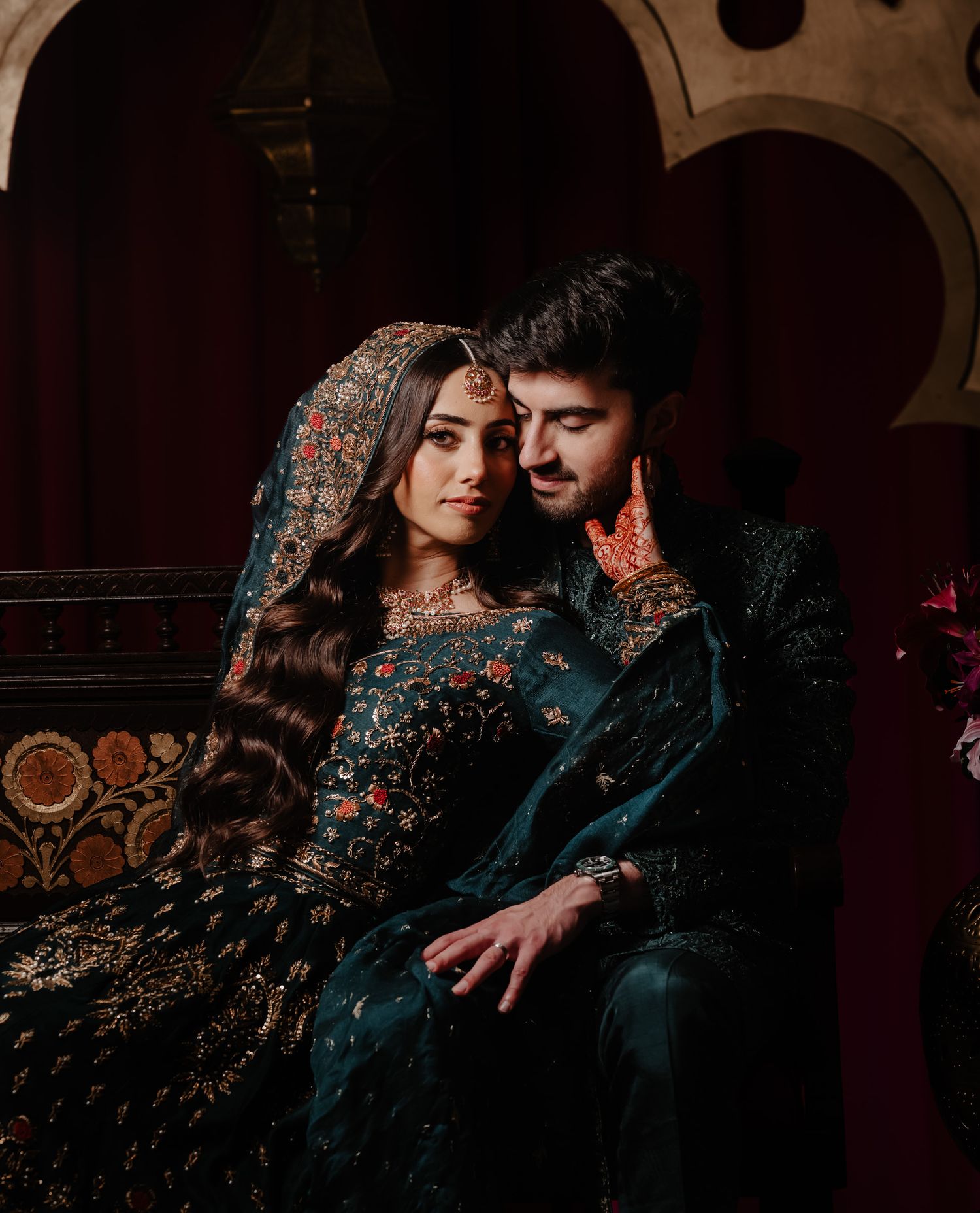 Romantic couple in traditional dark embroidered outfits pose intimately against arched architectural backdrop.