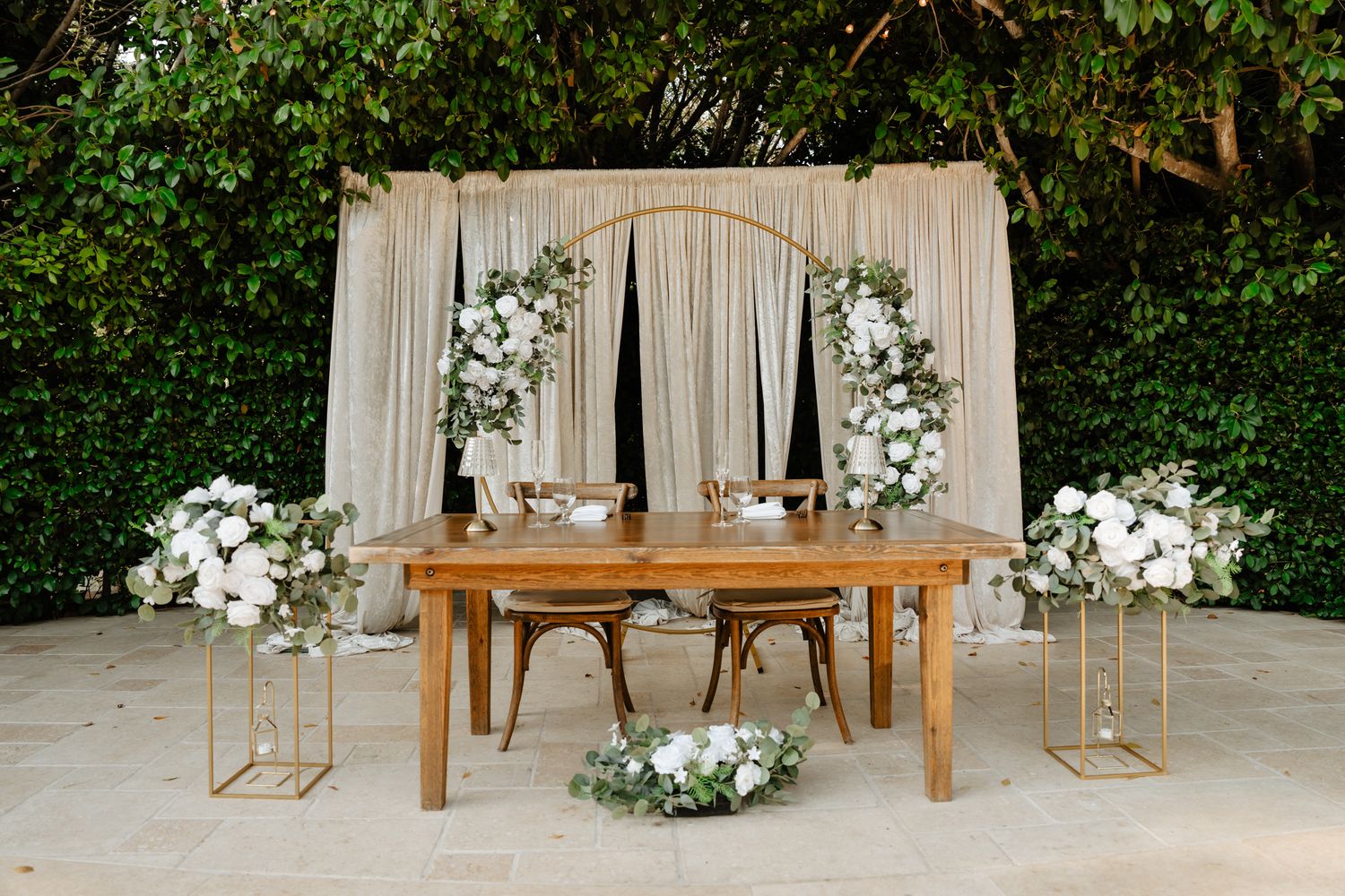 Elegant white wedding ceremony setup with floral arrangements and wooden benches against a green backdrop.