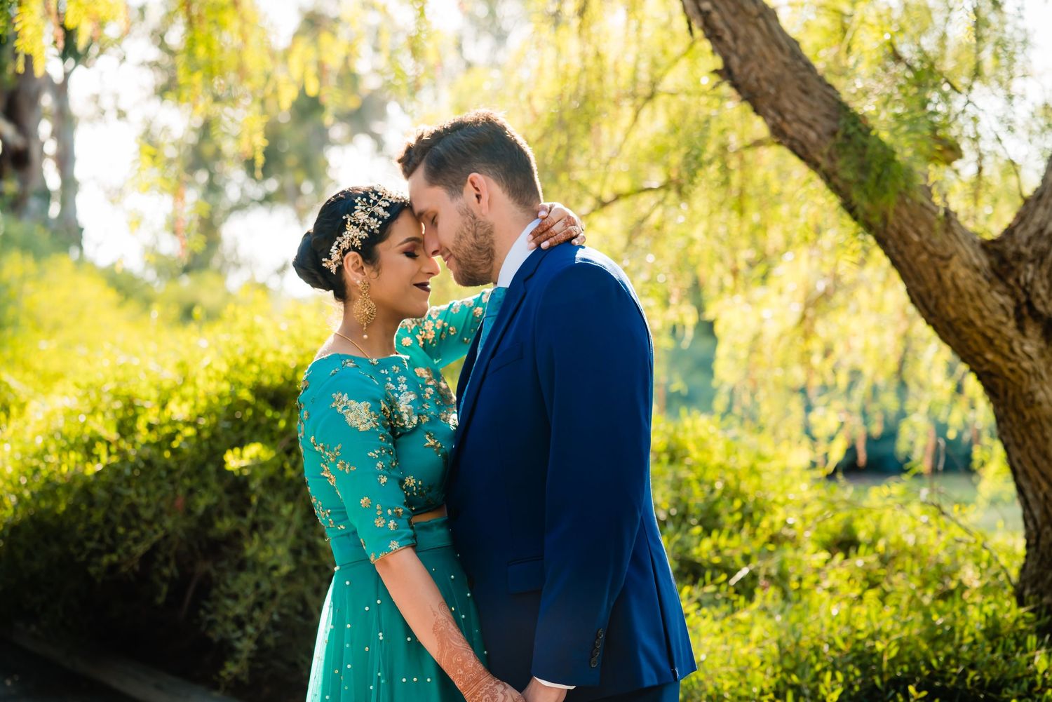 A couple in a turquoise dress and navy suit share romantic moments in a sunlit garden setting.