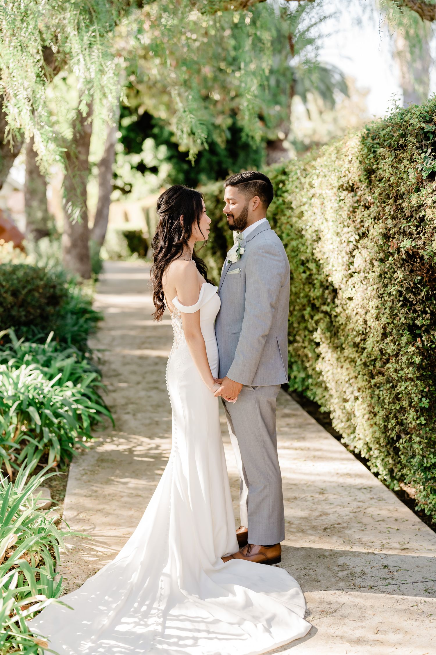 A couple in wedding attire stands together on a garden path surrounded by lush greenery.