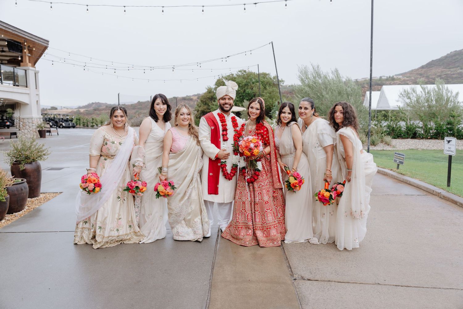 Indian wedding party poses outdoors, bride in red lehenga, groom in sherwani, bridesmaids in ivory sarees holding colorful bouquets.