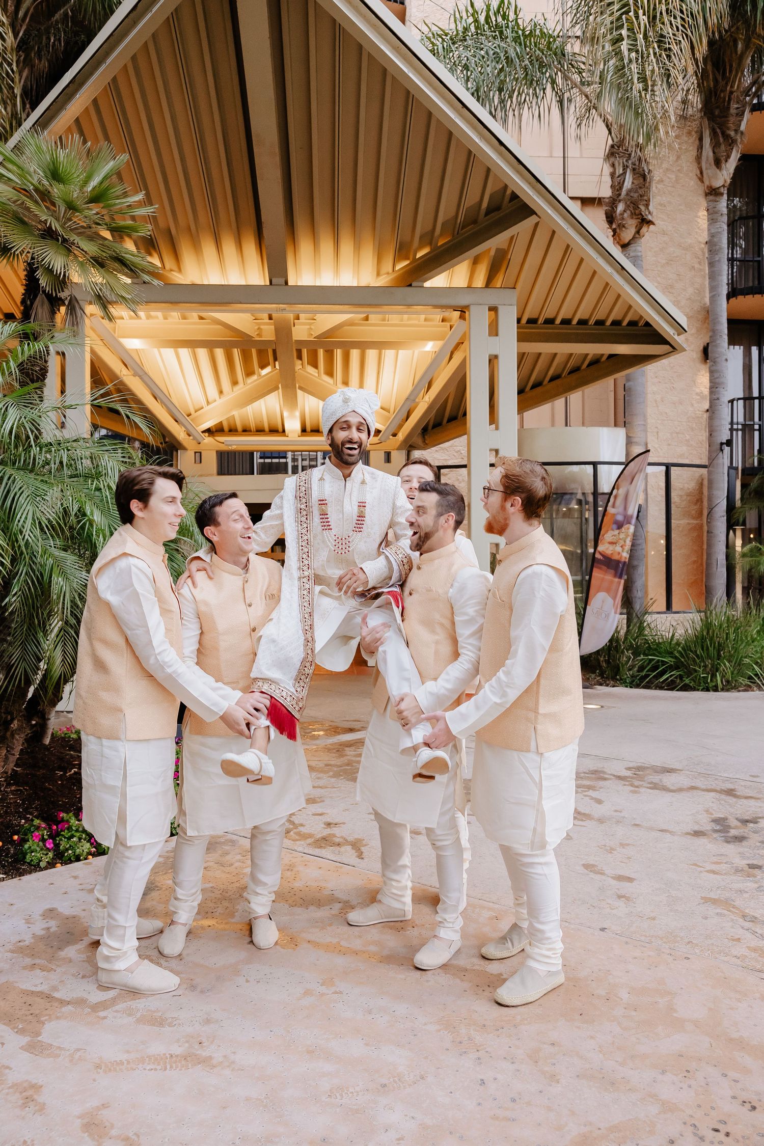 Groom in traditional Indian attire lifted by groomsmen in matching cream sherwanis outside a venue.