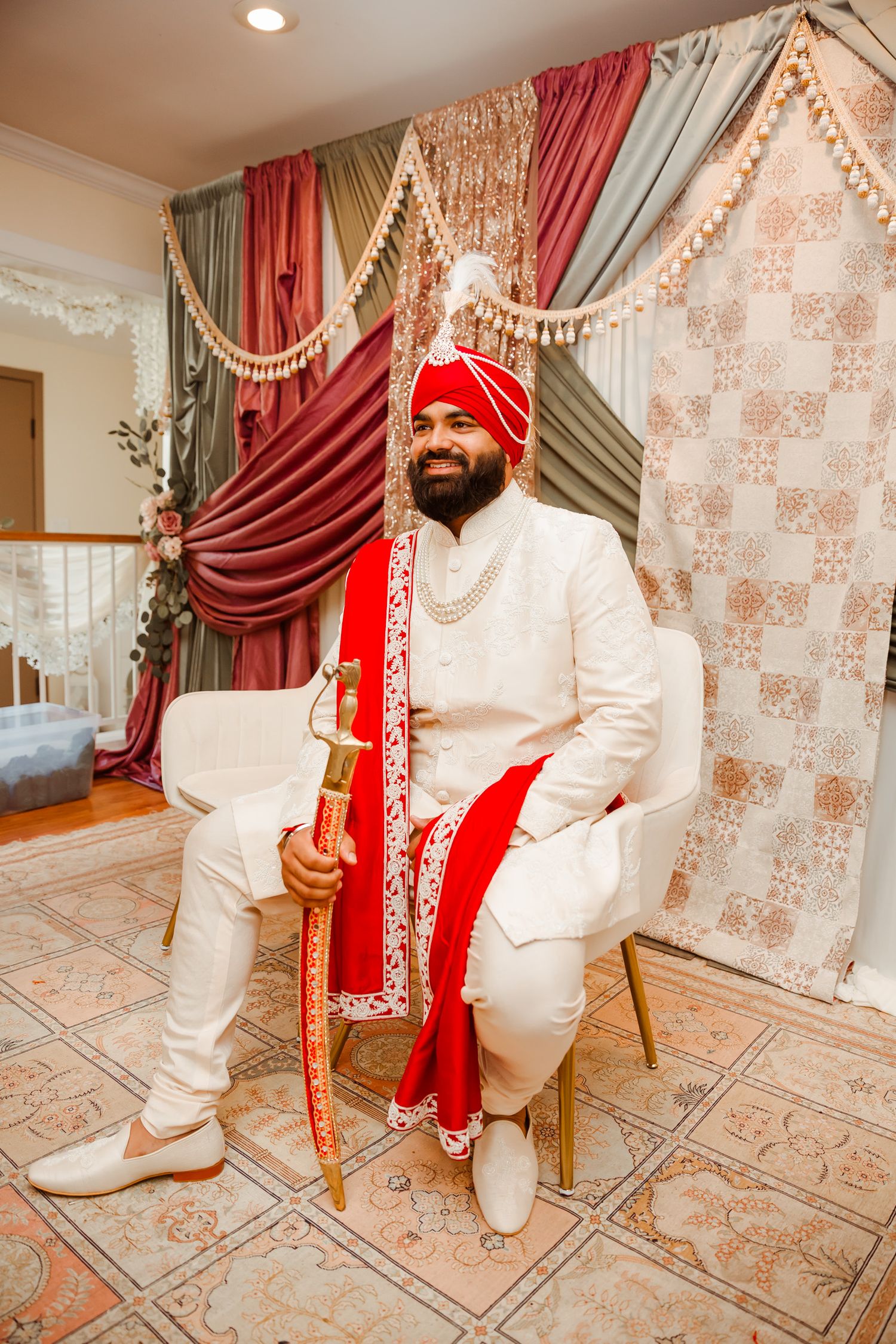 Traditional Indian groom in white sherwani and red turban poses in an elegantly decorated room with patterned carpet and drapes.