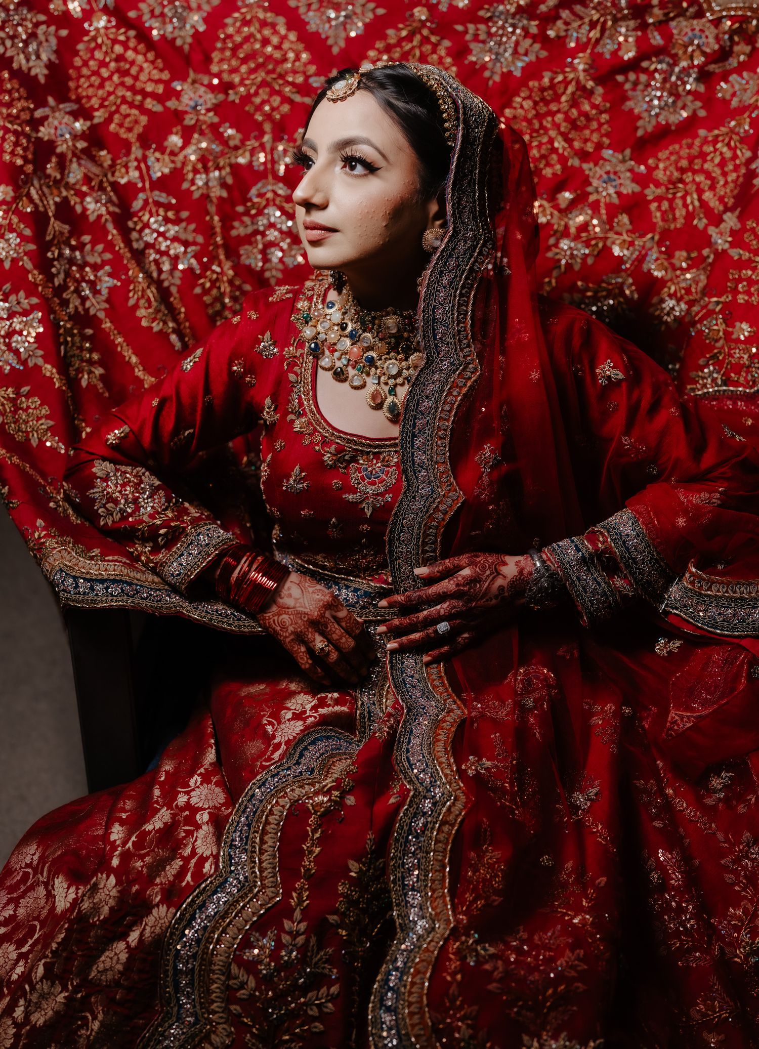 Traditional Indian bridal attire in deep red with intricate embroidery and jewelry against a red patterned backdrop.