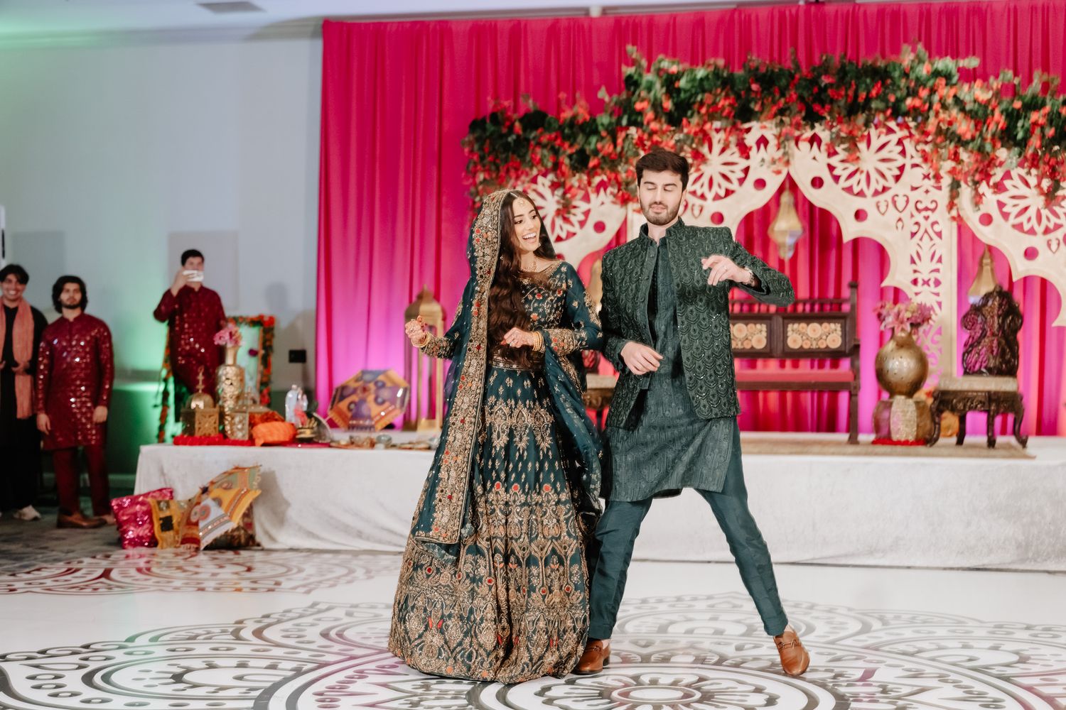 A couple performs a dance at an Indian wedding reception with a pink floral backdrop and decorative stage setup.