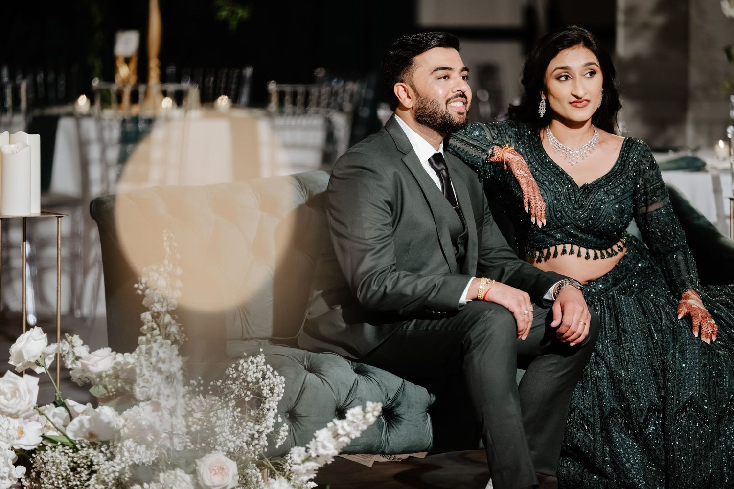 Couple in elegant dark green attire seated at a beautifully decorated wedding reception venue with floral arrangements.