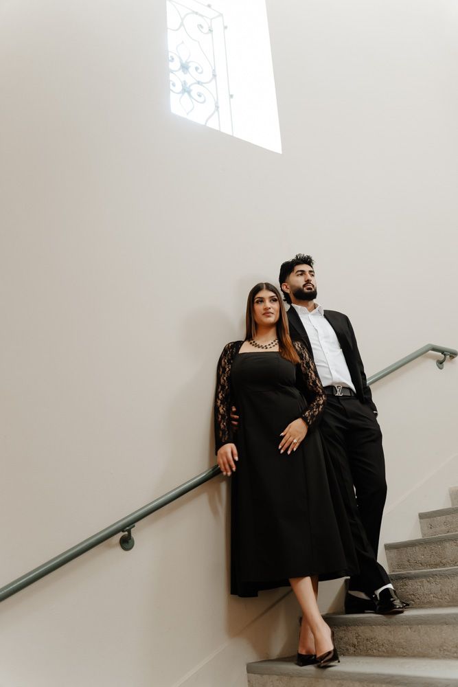 Elegant couple in matching black outfits posing on stairs against a white wall with decorative window above.