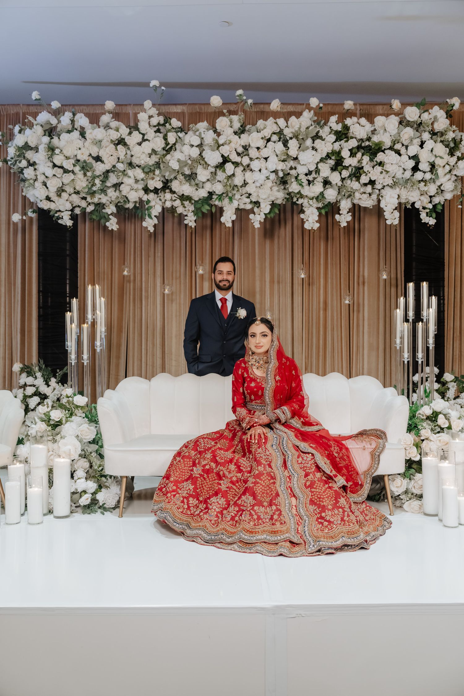 Wedding couple poses against white floral backdrop with elegant candles and modern decor elements.