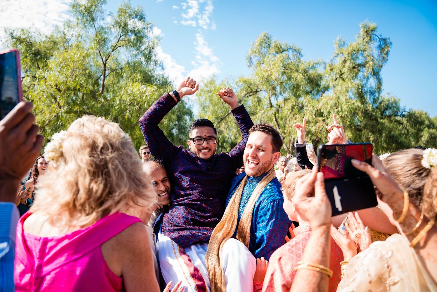 A group of people celebrate outdoors under blue sky and green trees during a festive gathering.