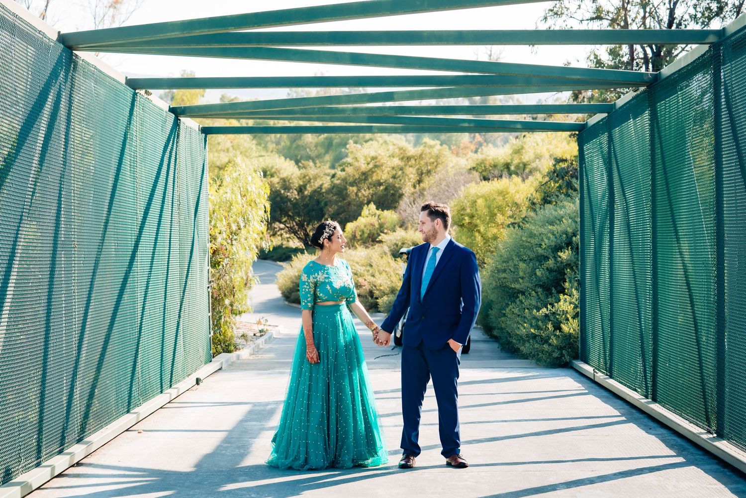 A couple stands on a turquoise-colored metal bridge walkway surrounded by green trees on a sunny day.