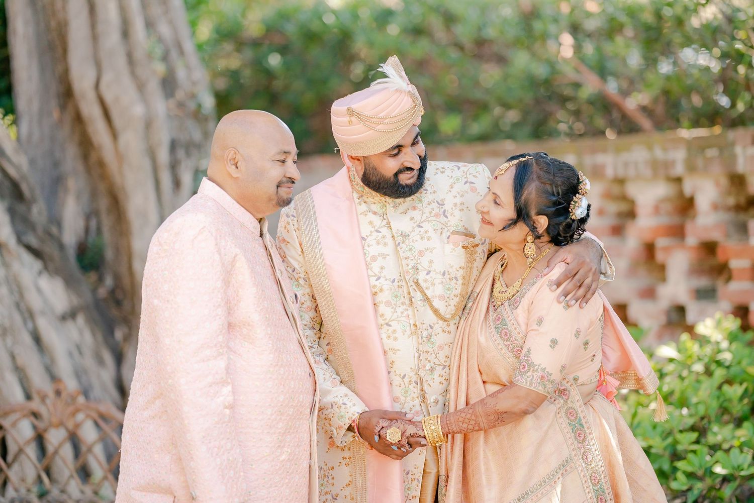 Three people wearing traditional South Asian wedding attire in soft pink and gold colors share a joyful moment in a garden setting.