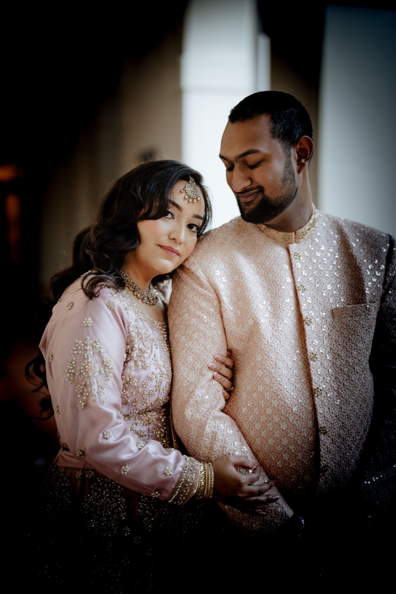 A couple in ornate South Asian formal wear share a tender moment in dramatic lighting.