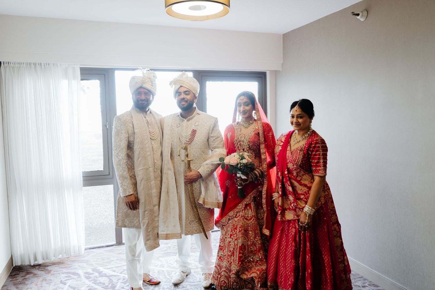 Four people in traditional Indian wedding attire pose together, with two men in sherwanis and two women in red lehengas.