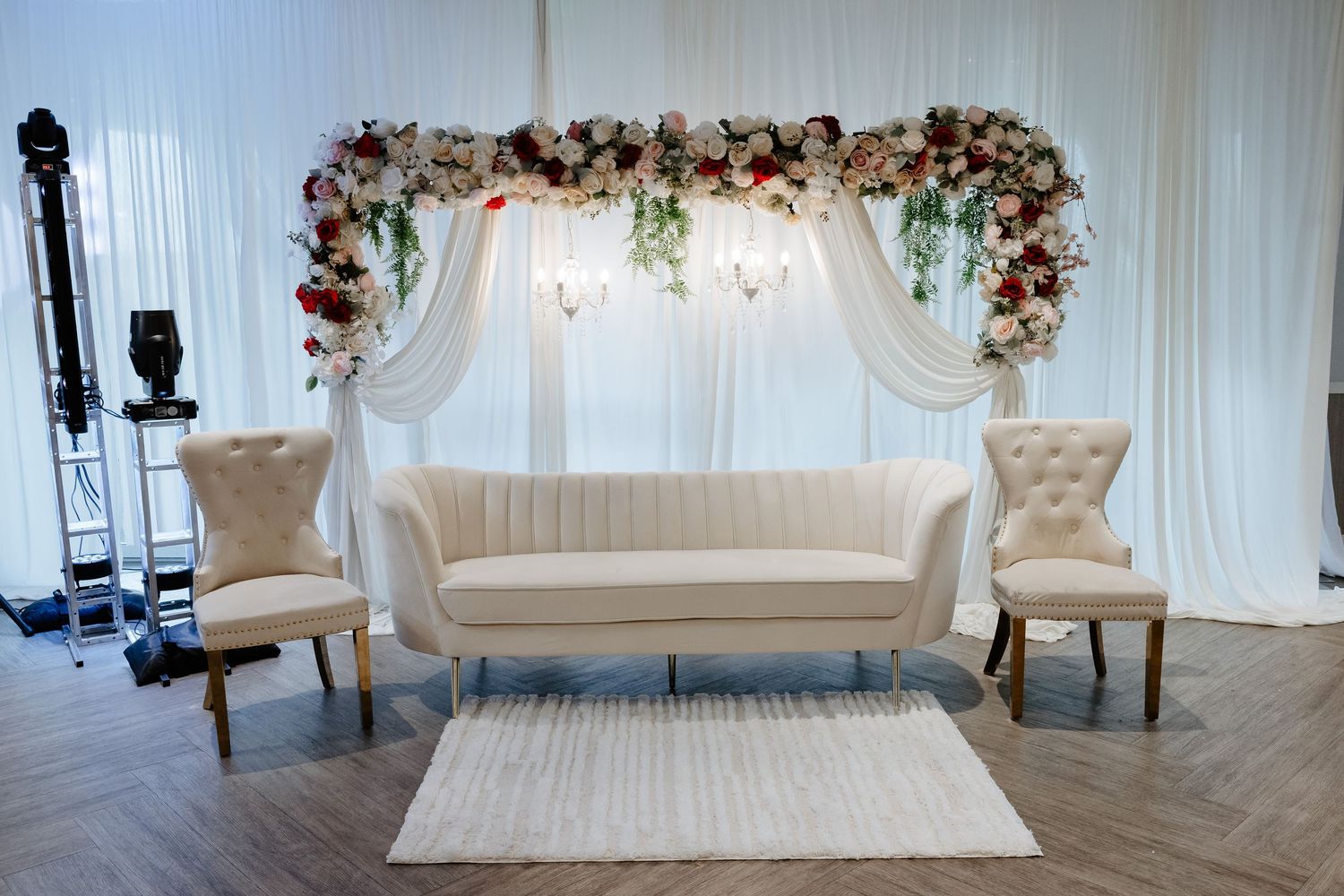 Elegant wedding sweetheart table setup with white sofa, tufted chairs, and floral arch backdrop with red and white blooms.
