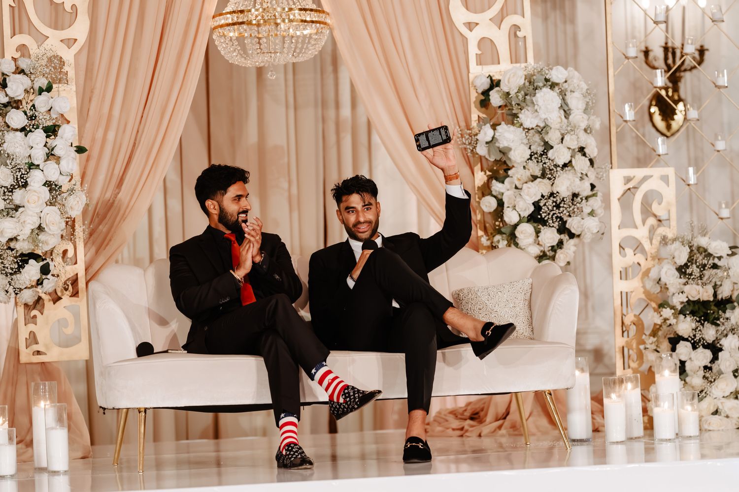 Two groomsmen in suits sitting on white couch with floral decor celebrating with fun poses.