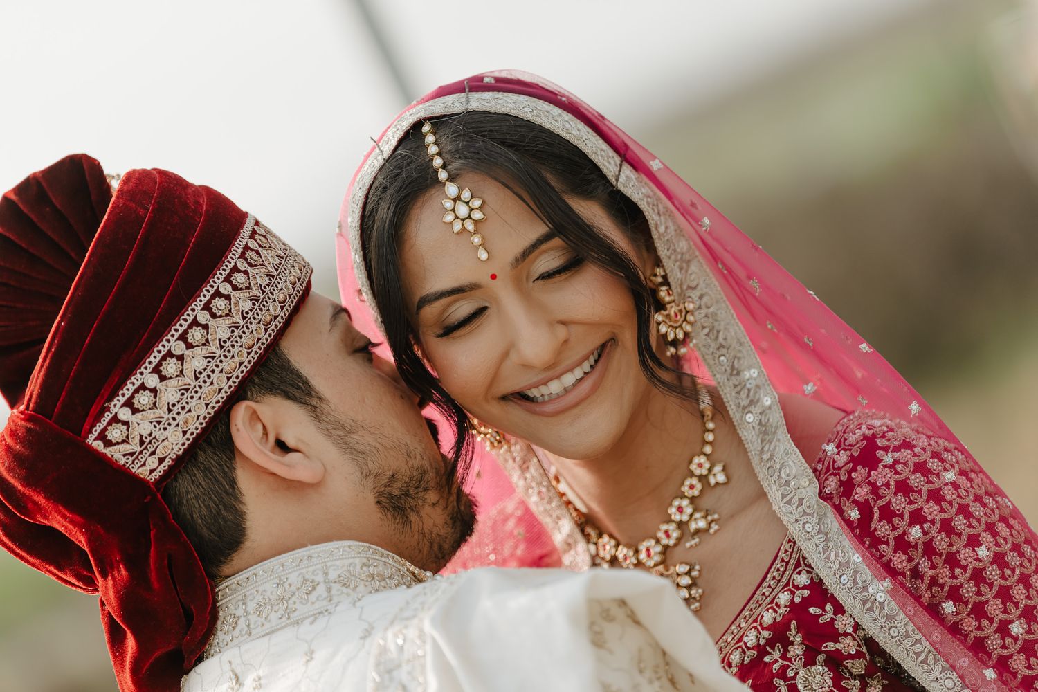 A romantic moment captured between a couple in traditional Indian wedding attire with red and pink colors.