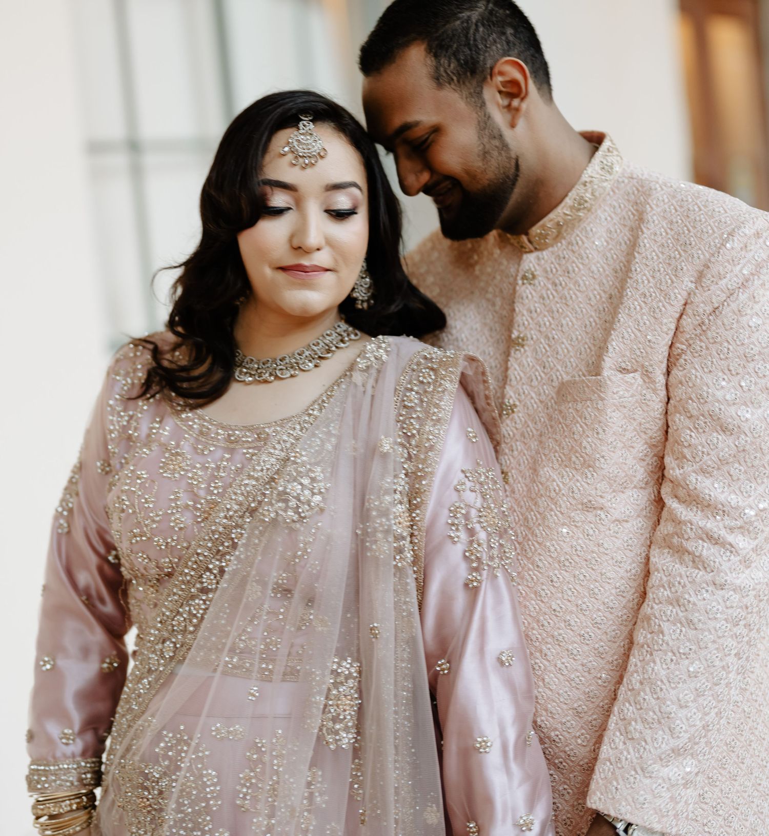 A romantic portrait of a couple in blush pink South Asian wedding outfits with intricate embroidery.