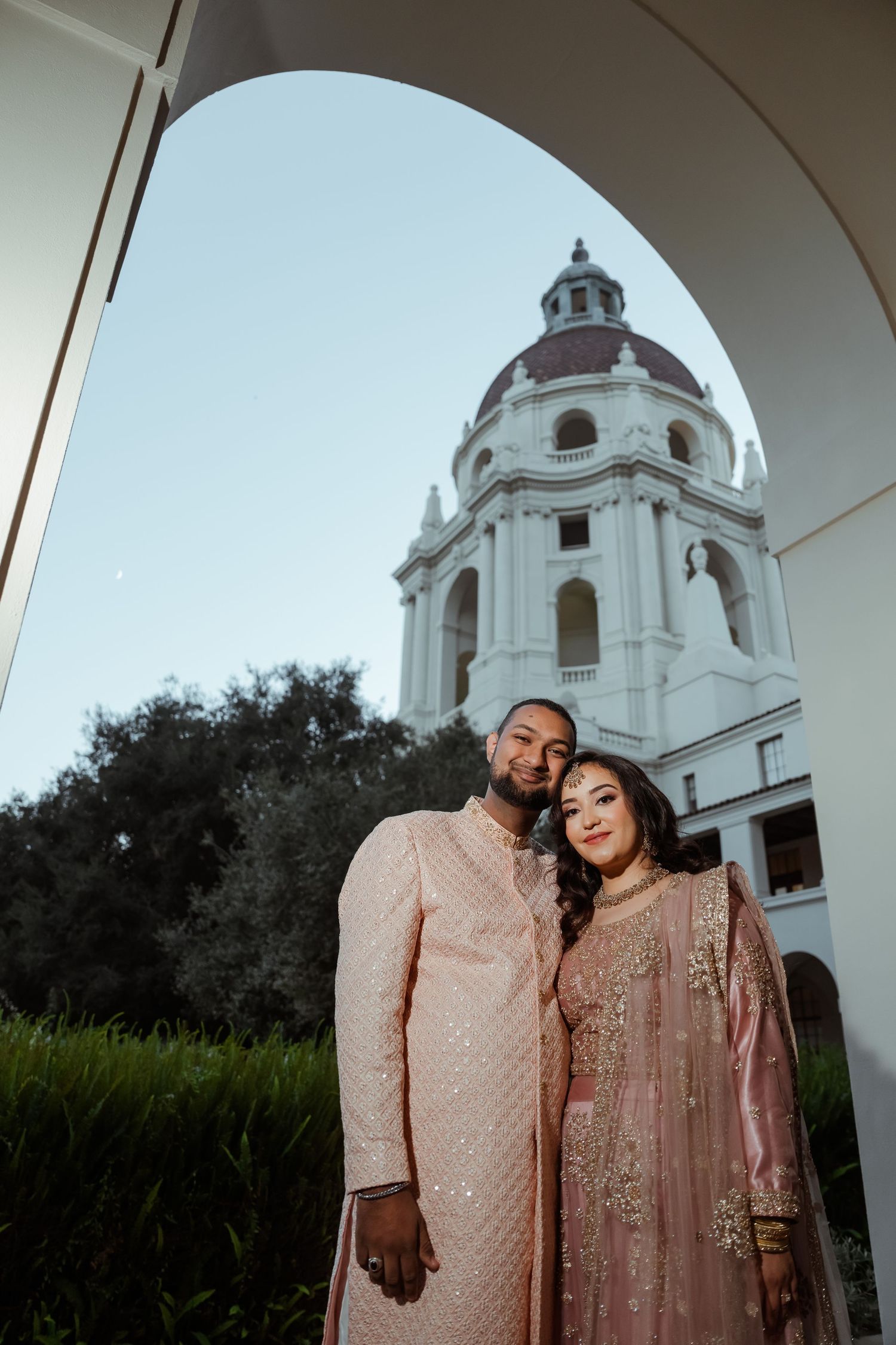 A couple in matching pink traditional outfits pose in front of a white domed building architecture.