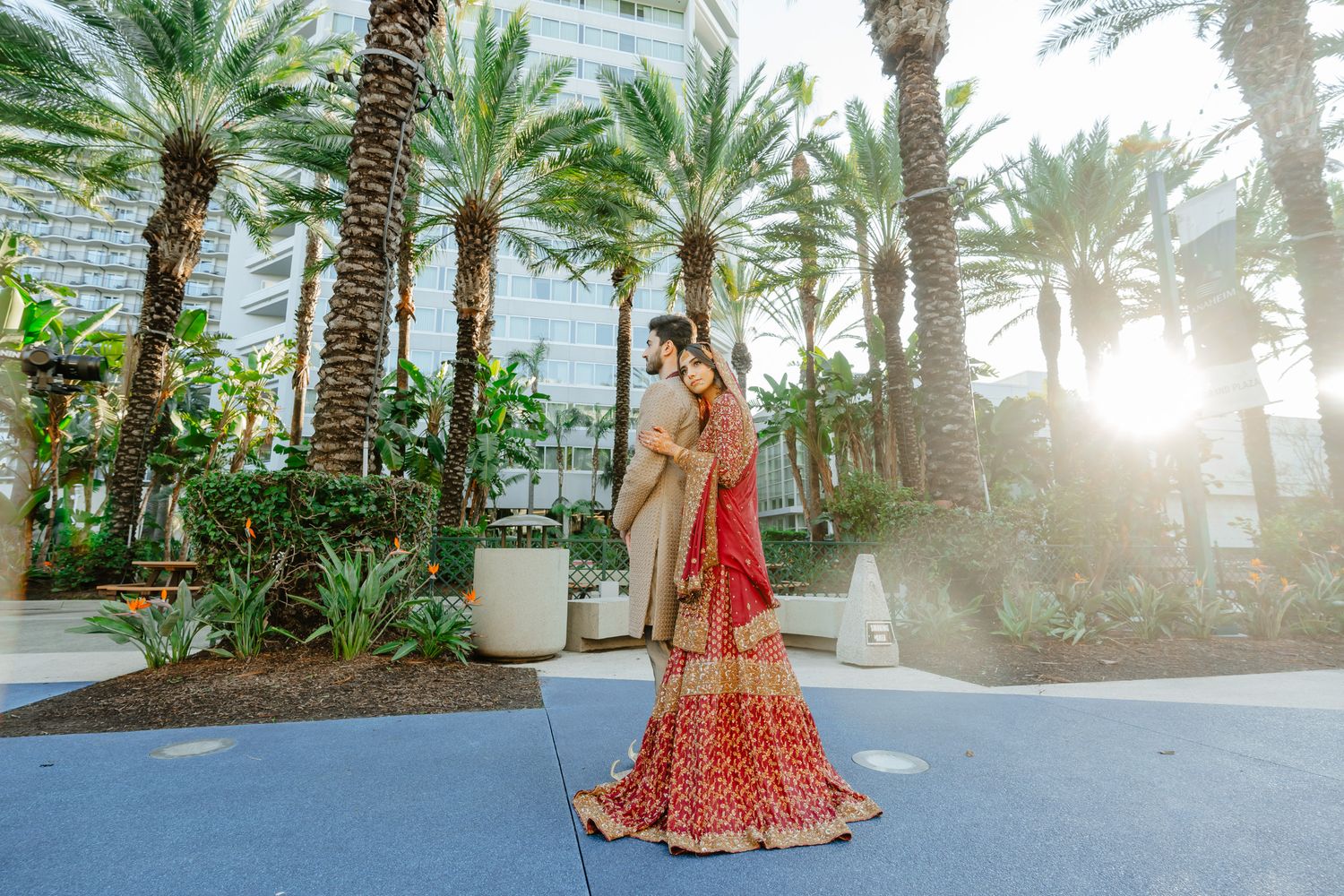 Traditional South Asian bride in red lehenga photographed among palm trees in golden sunlight.