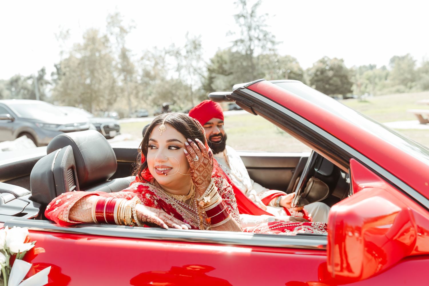 A couple in traditional Indian wedding attire pose in a red convertible car.
