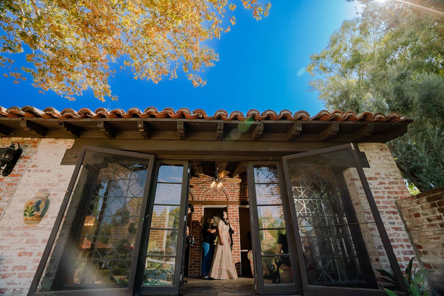A couple stands in the doorway of a rustic Spanish-style building with glass doors and autumn foliage overhead.
