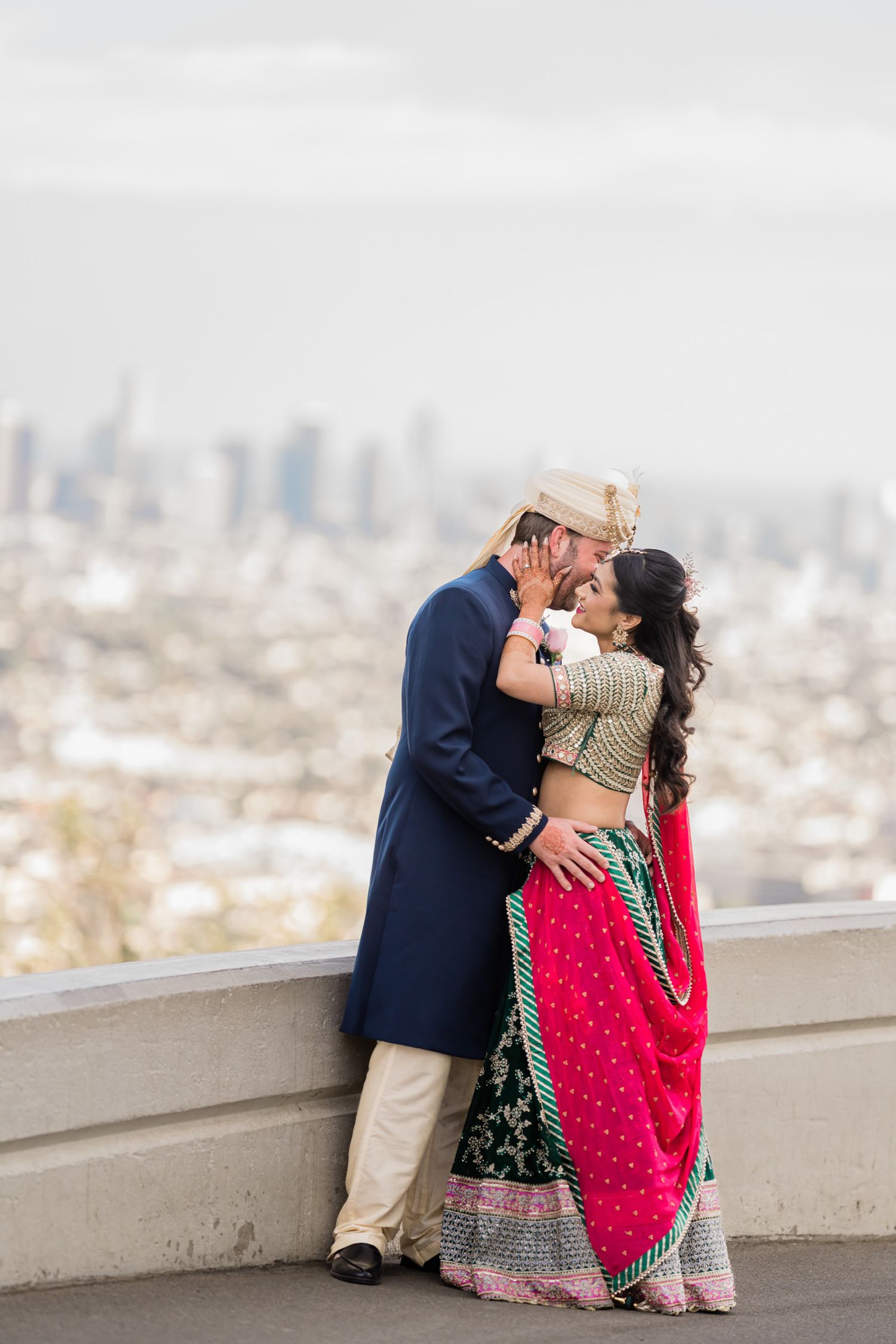 A couple in traditional South Asian attire embrace on a rooftop terrace with a cityscape backdrop.