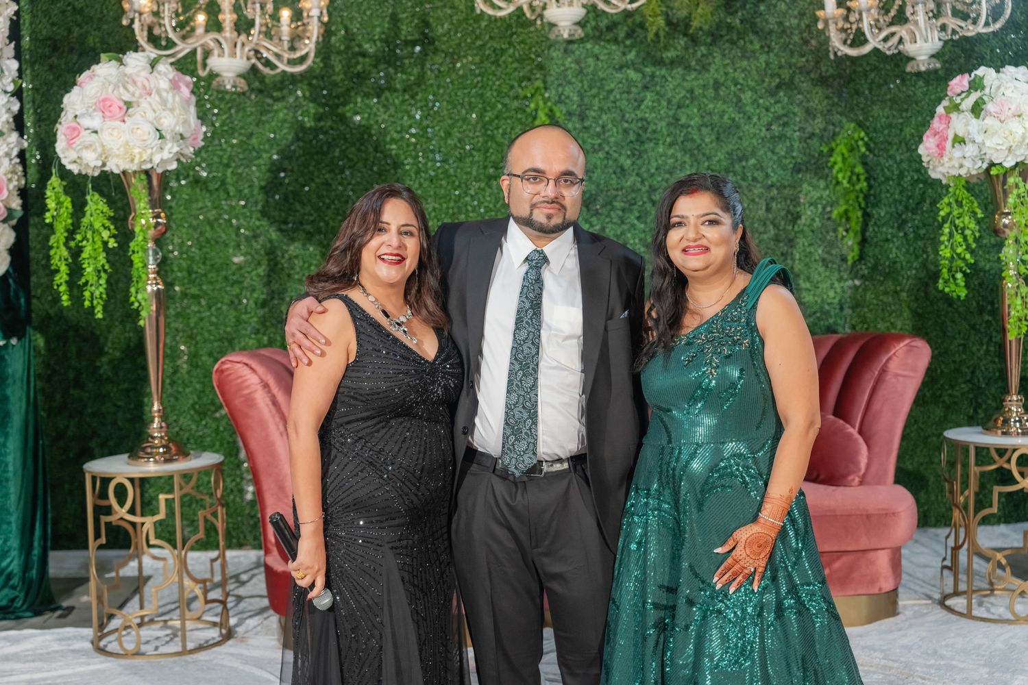 Wedding guests pose against a luxurious green hedge wall decorated with elegant chandeliers.