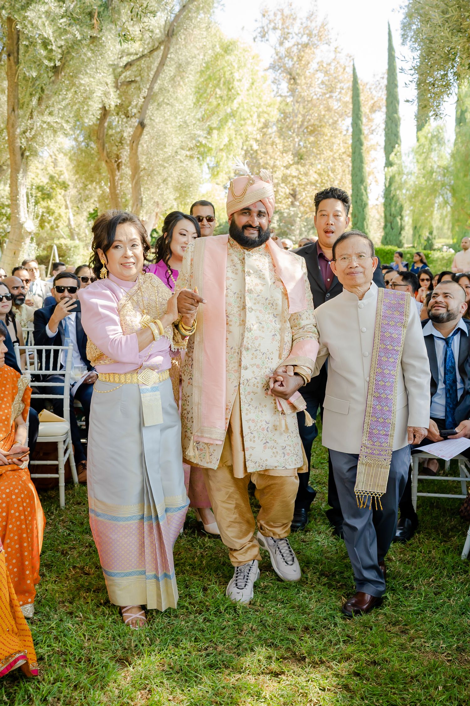 Large wedding party in traditional Indian and Western attire poses together outdoors under trees in natural lighting.