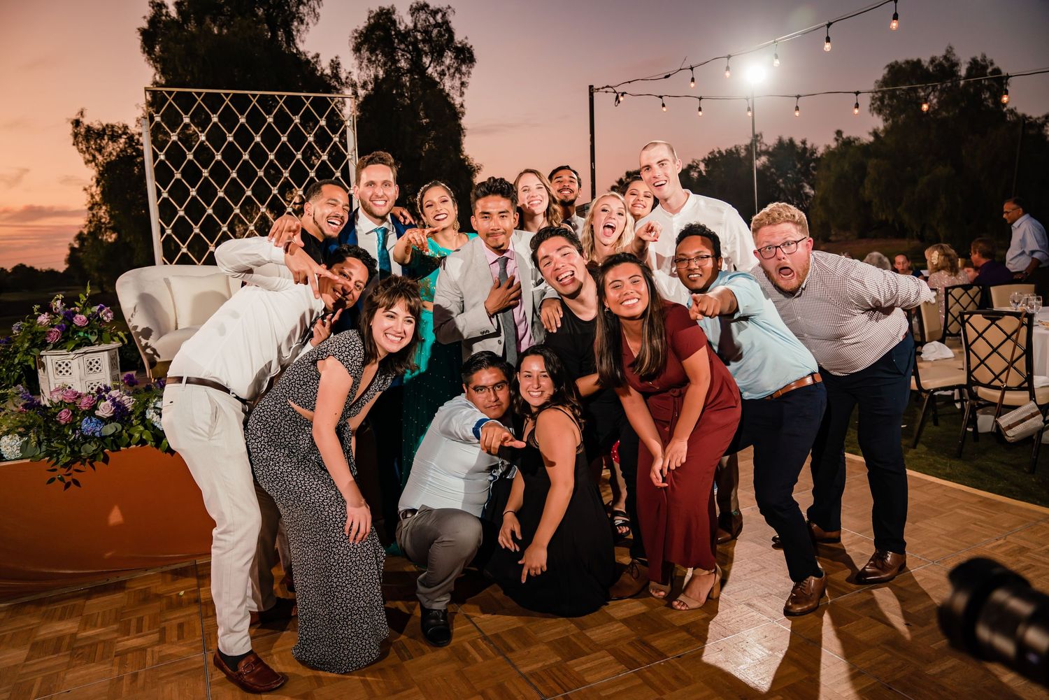 A group of wedding guests pose together on an outdoor dance floor at sunset with string lights overhead.